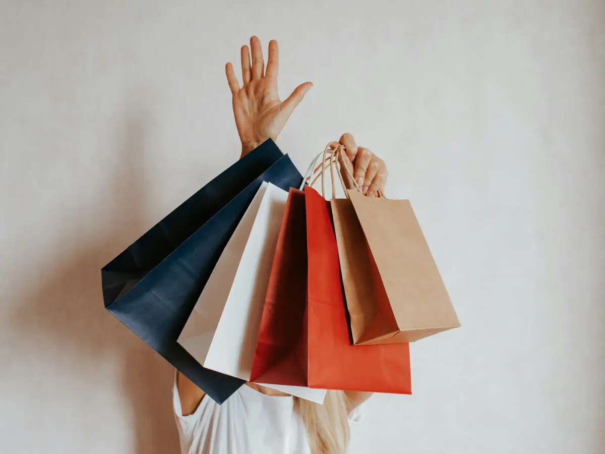 Un éventail de sacs de courses en papier bleu, blanc, rouge et kraft tenus en l’air devant un mur clair