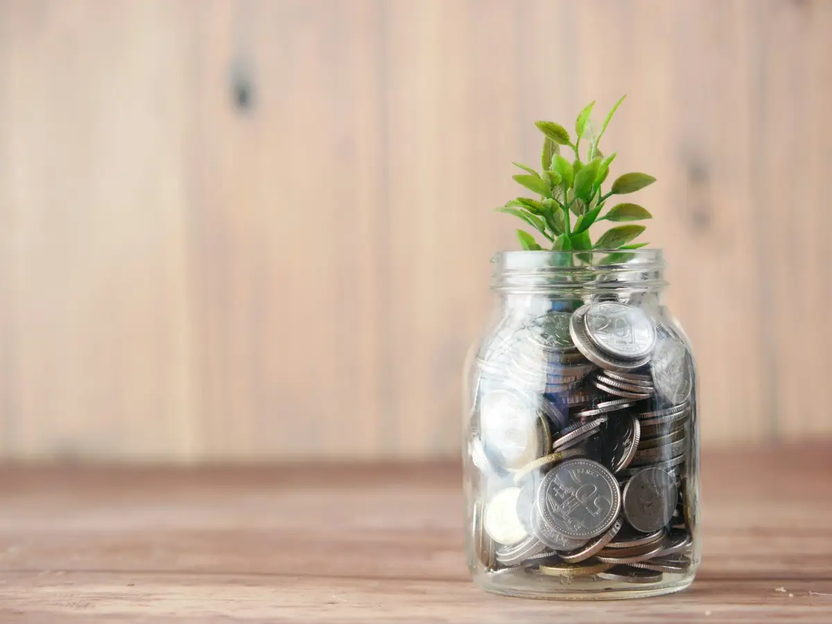 Un bocal en verre rempli de pièces de monnaie, surmonté d’une petite plante verte, posé sur une table en bois
